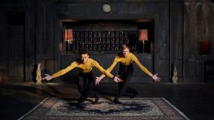 Two dancers in yellow long sleeved shirts and black pants are in a dark hotel lobby. Behind them is a worn black wall with cubbies, a clock, two mauve old fashioned table lights, and two small dog statues. The dancers are posed identically: bowed forward with their legs crossed and hands extended to their sides.