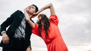 A dancer in a bright orange dress holds the head of another dancer, who is wearing a black suit. The dancer in the dress's long brown hair tumbles down around their face, while the other dancer looks into the sky, their light eyes matching the gray sky. Behind them is only sky.