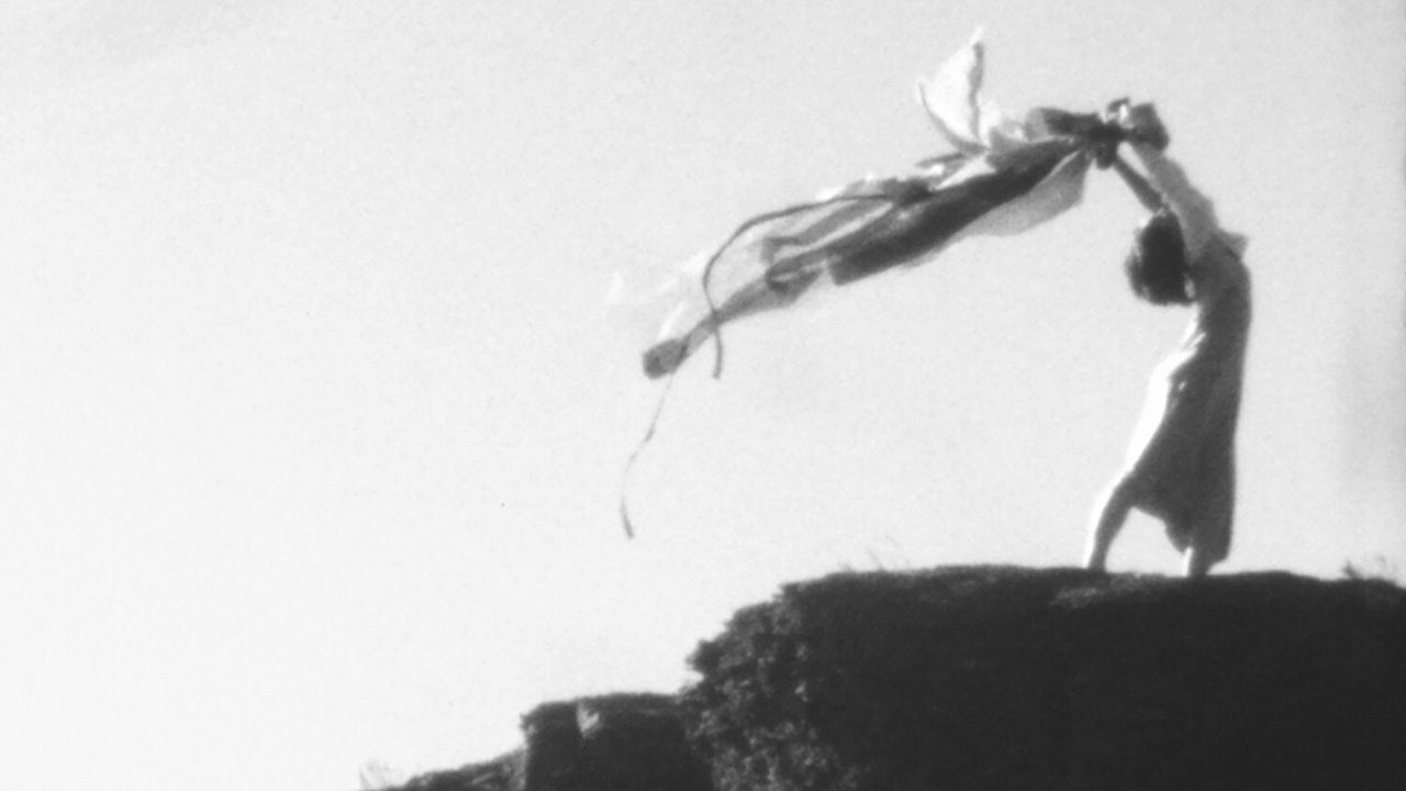 A grainy greyscale image of a dancer posing on a rocky hilltop. She wears a dress and arches backwards, holding a long piece of fabric above her head. The fabric blows behind her in the wind.