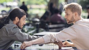 Two young men sit at outside a chess table, reaching across the board towards one another, as if for each other's pieces. They are holding deep, inquisitive eye contact.