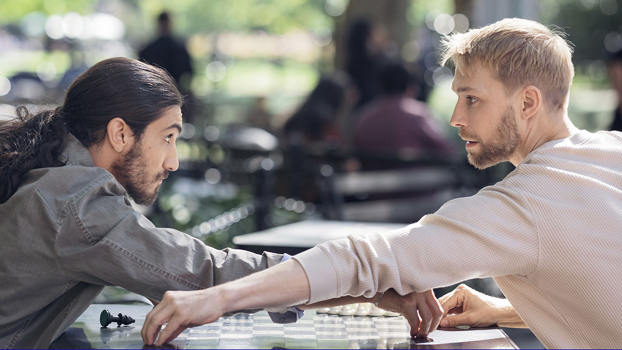 Two young men sit at outside a chess table, reaching across the board towards one another, as if for each other's pieces. They are holding deep, inquisitive eye contact.