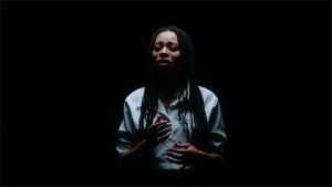 A singer emerges from a black background. She wears a crisp white button-up shirt. Her hands are slightly raised as she closes her eyes to sing.