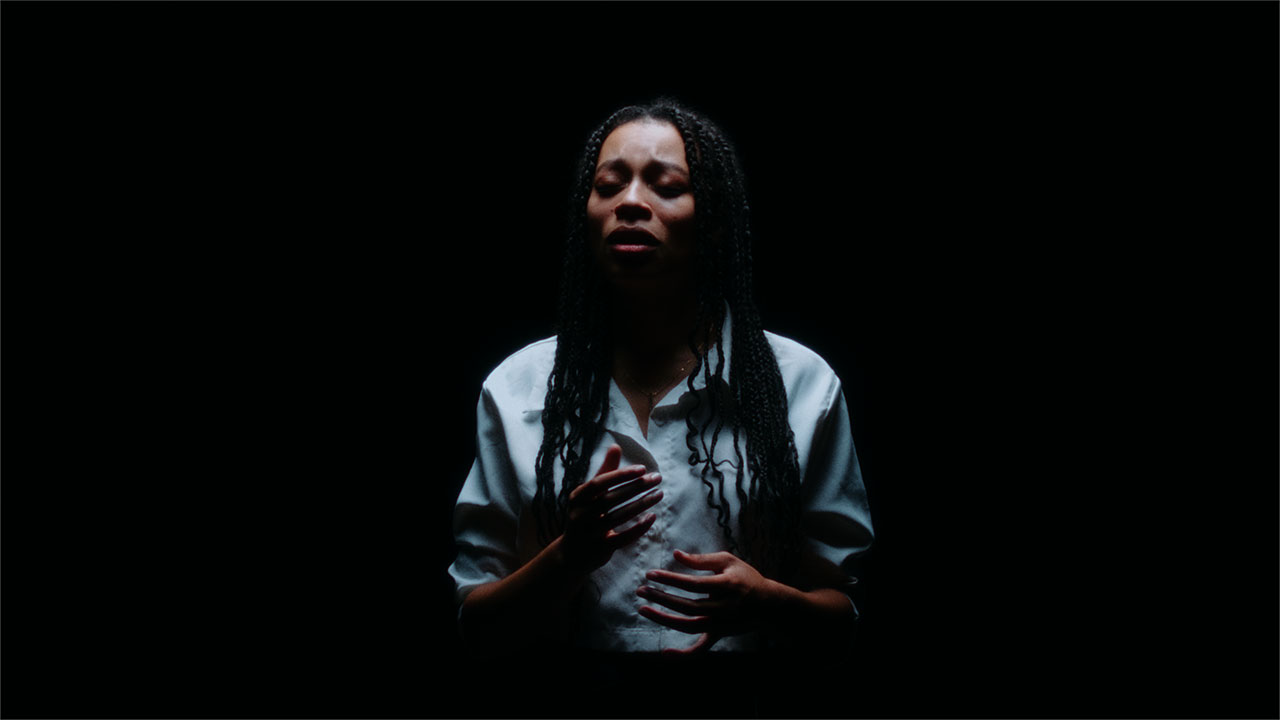 A singer emerges from a black background. She wears a crisp white button-up shirt. Her hands are slightly raised as she closes her eyes to sing.