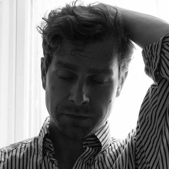 Jeremy has short light-colored hair and a scruffy beard and poses in front of a white sheer curtain with their upper left limb atop their head. Their eyes are closed and they wear a white striped dress shirt with the top button open.