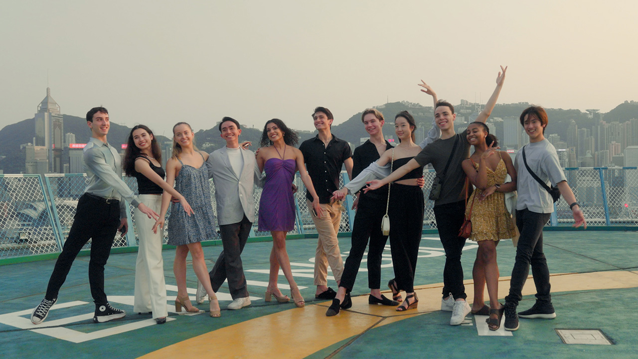 A group of nicely dressed high school-college age dancers pose for the camera with wide smiles. They all point a foot in front or behind themselves, and their arms are all up, pointed in front of them, or around each others' backs.