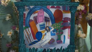 A colorful puppet show display, involing a dancer in an all white outfit and miscellaneous round shapes with various patterns (zigzags, stripes, dots, etc.). Behind the scene are real white and purple flowers.