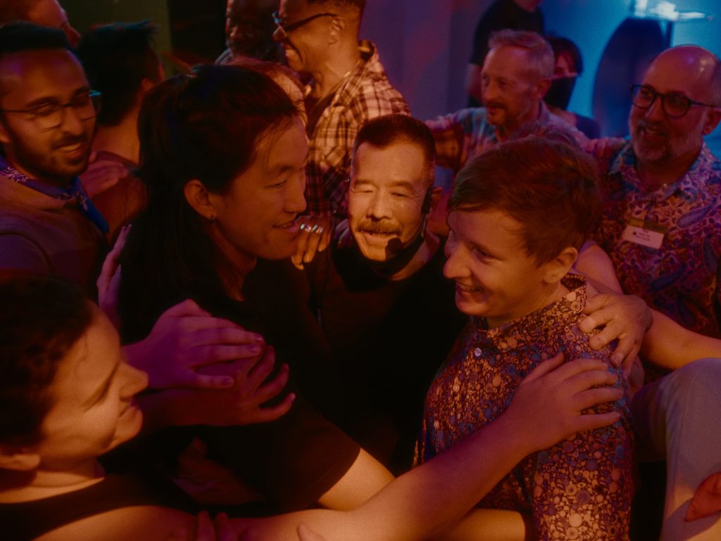 Sundance Stompede founder Ingu Yun gathers closely with participants in a joyful embrace. Individuals of diverse backgrounds place their hands gently on each other’s shoulders and backs, forming a tight-knit, intimate circle. The scene is bathed in warm lighting and the moment captures a sense of community, connection, and celebration.