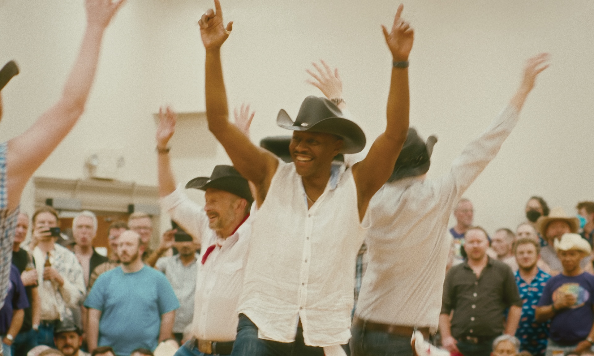 Film still from "Last Dance at the Sundance Stompede" A lively dance scene captures a diverse group of people in a western-themed setting. At the center, a man in a white sleeveless shirt and a black cowboy hat smiles broadly with his arms raised in the air, radiating joy.
