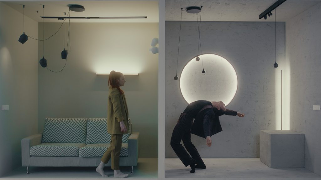The left half of the image shows a woman wearing a green suit in a small square room with a zigzag patterned couch. She is taking a step forward. On the right side, a man in a black suit leans far backwards, one arm reaching above his head and the other out to the side. Both rooms have lights on strings hanging from the ceiling.
