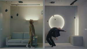 The left half of the image shows a woman wearing a green suit in a small square room with a zigzag patterned couch. She is taking a step forward. On the right side, a man in a black suit leans far backwards, one arm reaching above his head and the other out to the side. Both rooms have lights on strings hanging from the ceiling.