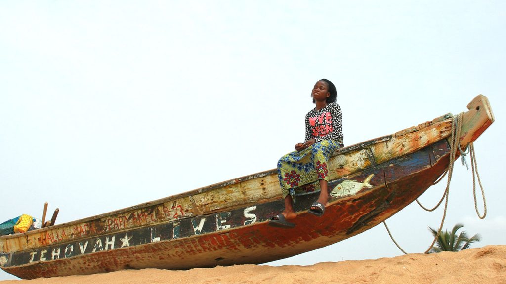 A girl sits on a boat in the sand in the bright sun, her legs dangling off the edge of the boat. She is wearing a brightly patterned shirt and skirt, and black slides that are hanging off her feet. She looks into the distance, her face contemplative.