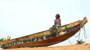 A girl sits on a boat in the sand in the bright sun, her legs dangling off the edge of the boat. She is wearing a brightly patterned shirt and skirt, and black slides that are hanging off her feet. She looks into the distance, her face contemplative.