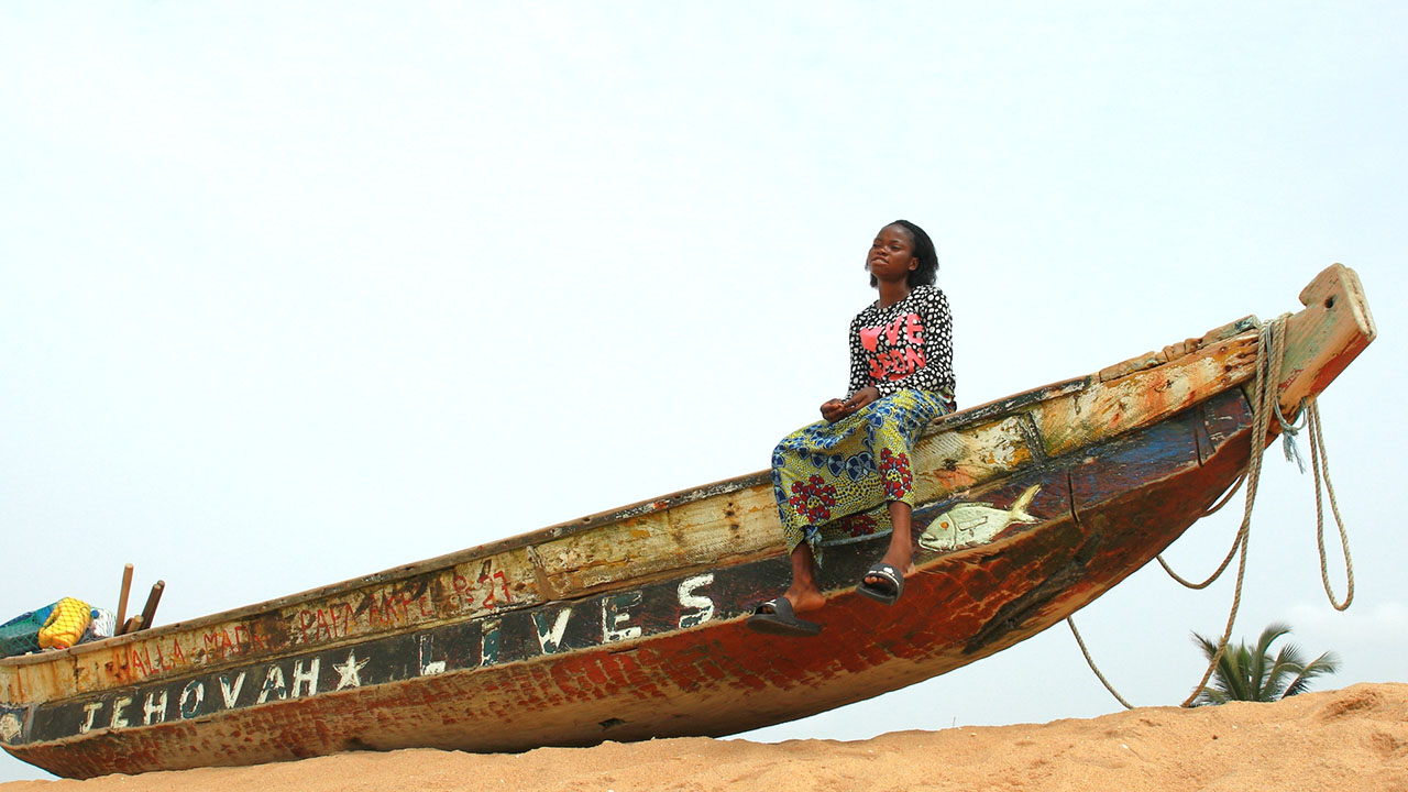 A girl sits on a boat in the sand in the bright sun, her legs dangling off the edge of the boat. She is wearing a brightly patterned shirt and skirt, and black slides that are hanging off her feet. She looks into the distance, her face contemplative.