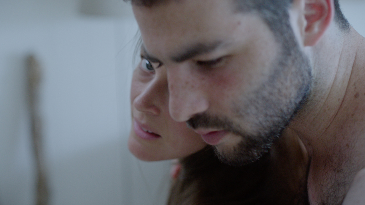 A woman's face pokes out from behind a man's face as she stands with her back against his bare chest. She looks directly at the camera, while he looks down, eyebrows furrowed. Image is slightly out of focus.