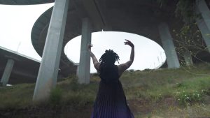 A black dancer in a long dress faces upwards and away from the camera, their arms gently reaching upwards. They stand in the grass under a concrete overpass.