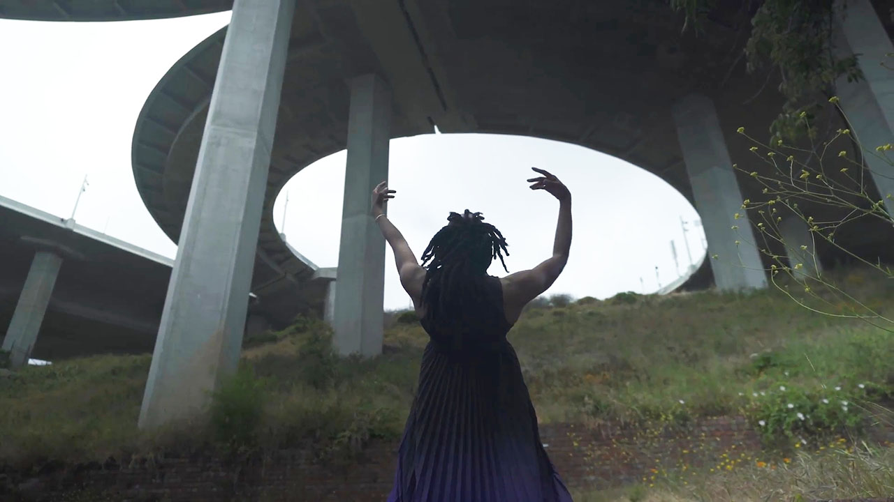 A black dancer in a long dress faces upwards and away from the camera, their arms gently reaching upwards. They stand in the grass under a concrete overpass.