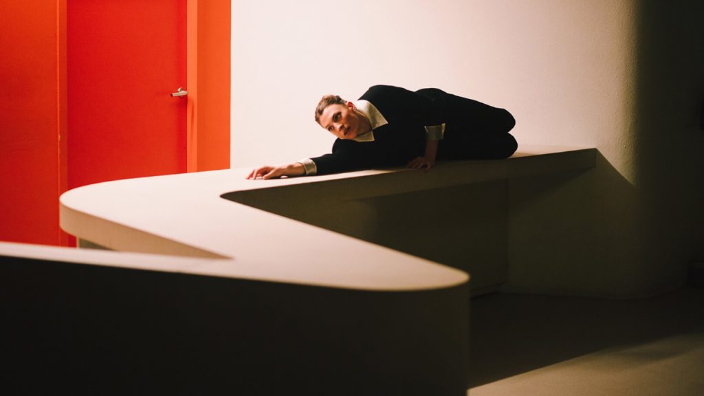 A woman lies along an s-shaped ledge, one arm outstretched as she looks down the length of the ledge. Behind her is a bright orange door.