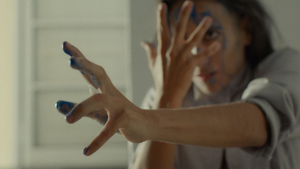 A dancer looks intensely forward with blue paint dripping from their hands and face. One hand is held to their face, fingers splayed, while the other is outstreched forward with tension.