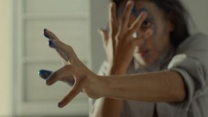 A dancer looks intensely forward with blue paint dripping from their hands and face. One hand is held to their face, fingers splayed, while the other is outstreched forward with tension.