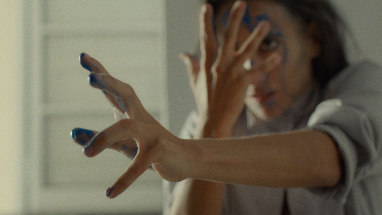 A dancer looks intensely forward with blue paint dripping from their hands and face. One hand is held to their face, fingers splayed, while the other is outstreched forward with tension.