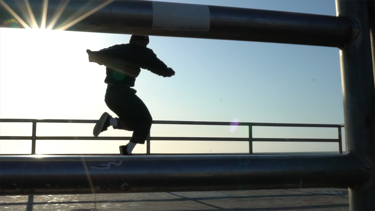 A dancer wearing every day street clothes and sneakers spins in the air, their jacket twirling behind them. The sun shines into the camera, creating a star-shaped flare. A metal railing obstructs the view of their feet in the foreground.