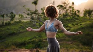 A woman with curly brown and bonde hair extends her arms out to her sides. The sun lights up the edges of her hair. She wears a blue, patterned bra and gray flowy pants. In the background is lush greenery.