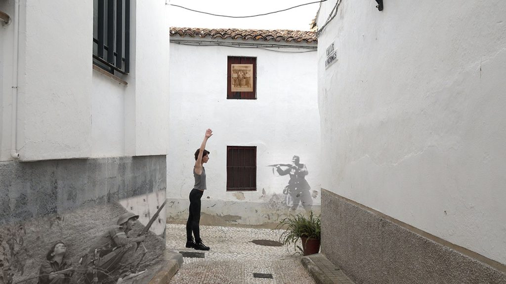 A woman in modern-day clothing stands in an alley with her arms up, facing a spray-painted image of an early 20th Century soldier on a wall. The soldier holds up a gun pointed in her direction.