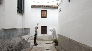 A woman in modern-day clothing stands in an alley with her arms up, facing a spray-painted image of an early 20th Century soldier on a wall. The soldier holds up a gun pointed in her direction.