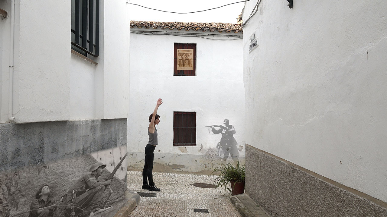 A woman in modern-day clothing stands in an alley with her arms up, facing a spray-painted image of an early 20th Century soldier on a wall. The soldier holds up a gun pointed in her direction.