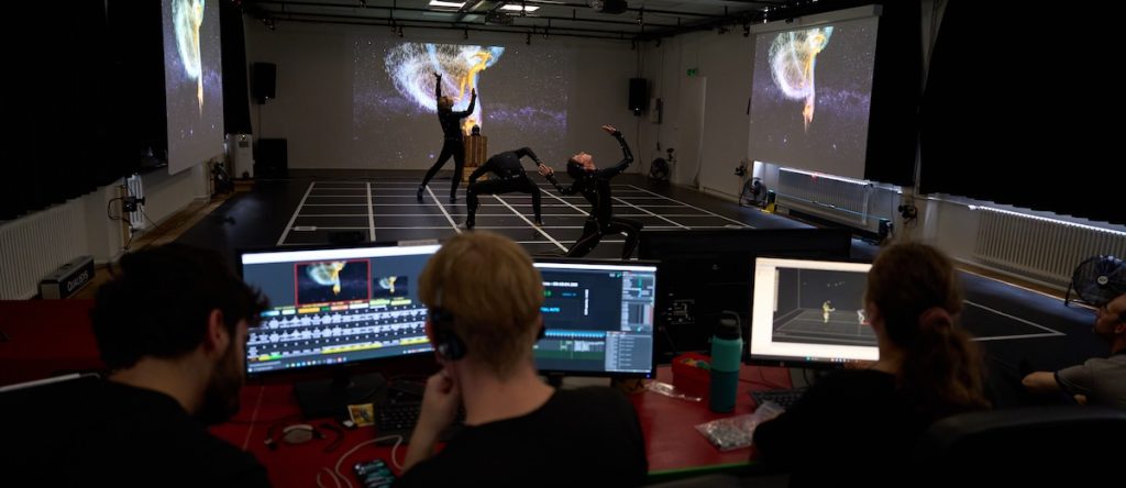 In the foreground, people sit in front of computer monitors running a motion-capture program. In the background, dancers wearing black move atop a white grid with projections around them.