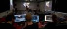 In the foreground, people sit in front of computer monitors running a motion-capture program. In the background, dancers wearing black move atop a white grid with projections around them.