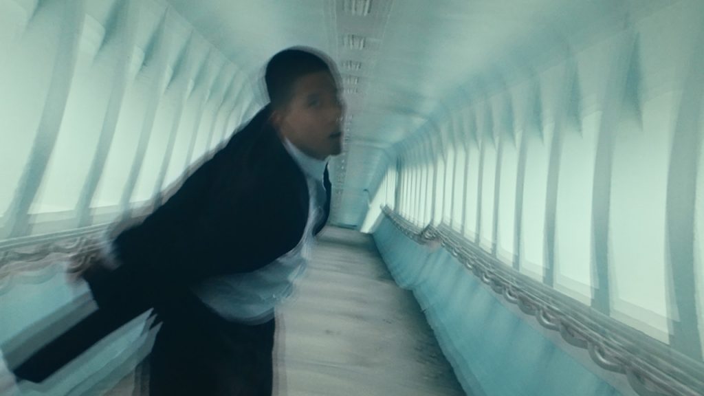 A blurred image of a tuxedo-wearing Asian dancer moving through a tunnel with light blue walls. The image feels unbalanced and the dancer glances at the camera with his mouth slightly open.