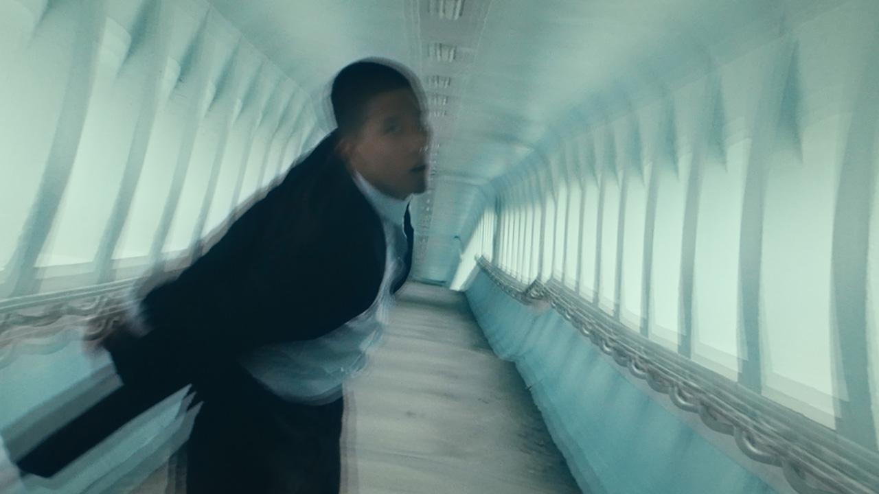 A blurred image of a tuxedo-wearing Asian dancer moving through a tunnel with light blue walls. The image feels unbalanced and the dancer glances at the camera with his mouth slightly open.