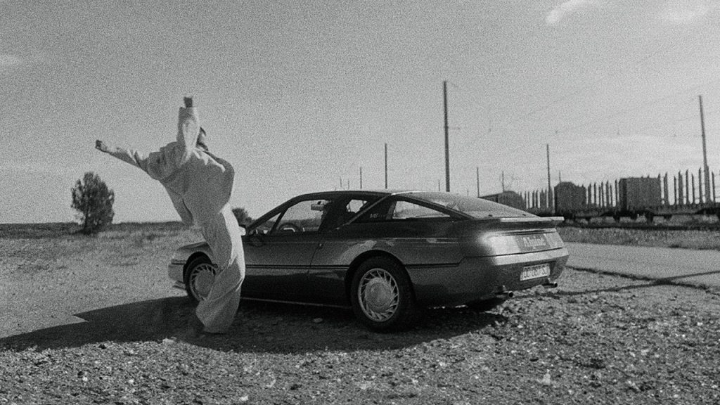 A person wearing an all-white outfit reaches their arms with balled fists upwards beside a parked car on a deserted gravel road with railway tracks in the background. The image is in black and white.
