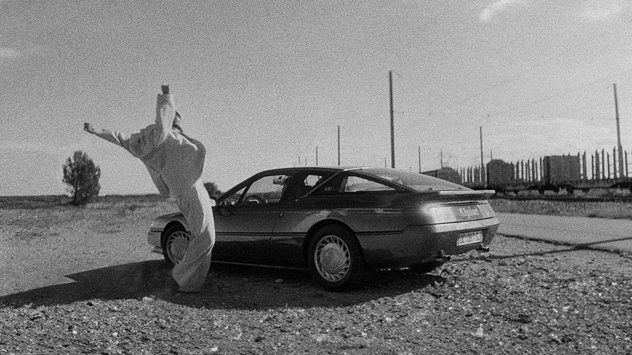 A person wearing an all-white outfit reaches their arms with balled fists upwards beside a parked car on a deserted gravel road with railway tracks in the background. The image is in black and white.