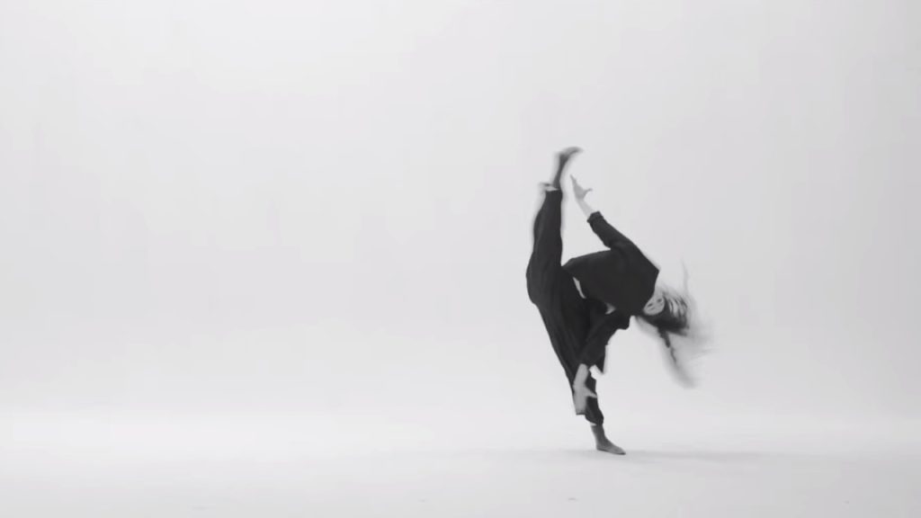 A blurry greyscale photo of a dancer wearing all black. They tip sideways, lifting one leg into a full side split, their hair wild.