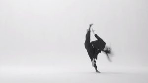 A blurry greyscale photo of a dancer wearing all black. They tip sideways, lifting one leg into a full side split, their hair wild.