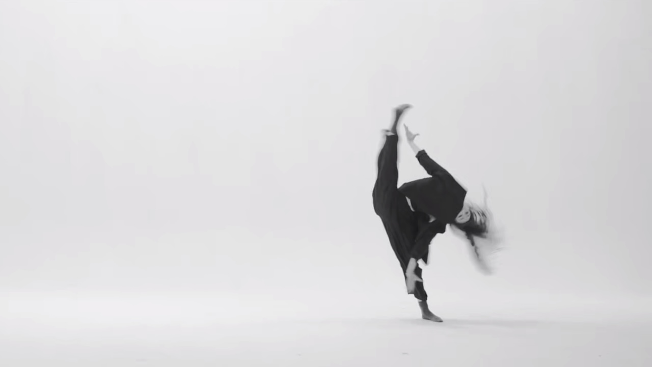 A blurry greyscale photo of a dancer wearing all black. They tip sideways, lifting one leg into a full side split, their hair wild.