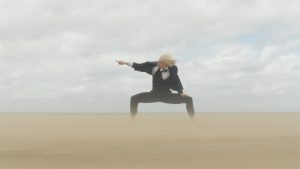 A person in a black and white suit stands on a sandy beach in a center lunge, both the sand and their hair blowing wildly in the wind. The dancer is pointing a finger out to the side, into the wind.