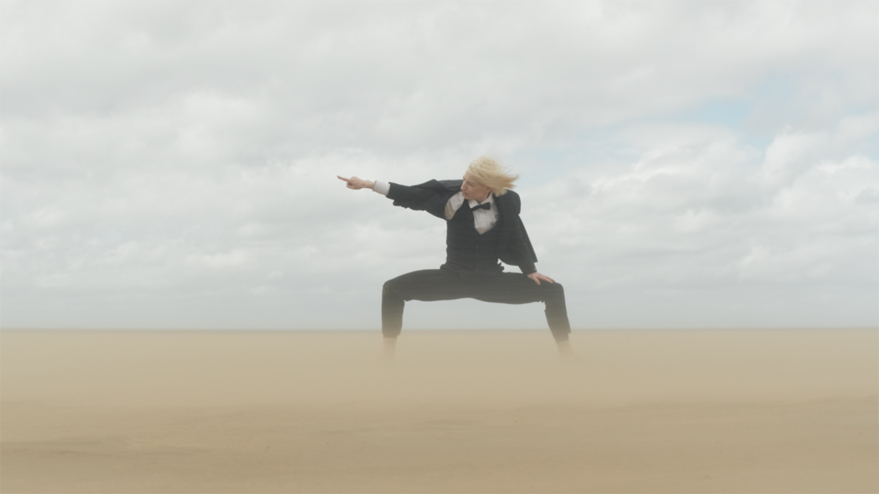 A person in a black and white suit stands on a sandy beach in a center lunge, both the sand and their hair blowing wildly in the wind. The dancer is pointing a finger out to the side, into the wind.