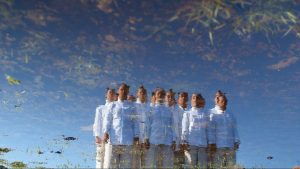 A reflection in a shallow body of water shows a group of dancers in white standing with their arms at their sides. The sky behind them is completely clear of clouds.