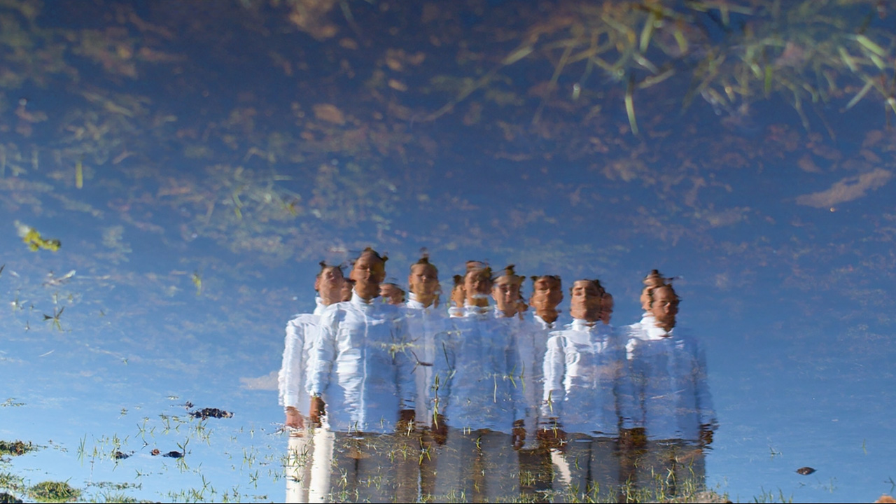 A reflection in a shallow body of water shows a group of dancers in white standing with their arms at their sides. The sky behind them is completely clear of clouds.