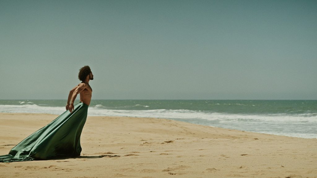A dancer stands on a beach, looking towards the ocean. They are shirtless, wearing a silk, green skirt that extends far behind them. They hold their arms back behind them, with their elbows backwards and chest out.