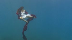 An underwater picture of a person in a flowing white dress holding a long red scarf that extends far below them in the water.
