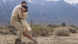 Two dancers wear beige colored outfits. One dancer hangs on the shoulders of the other in a hug, her legs just touching the ground behind her. She wears blue makeup in a line across her eyes. There are snow-capped mountains in the distance.