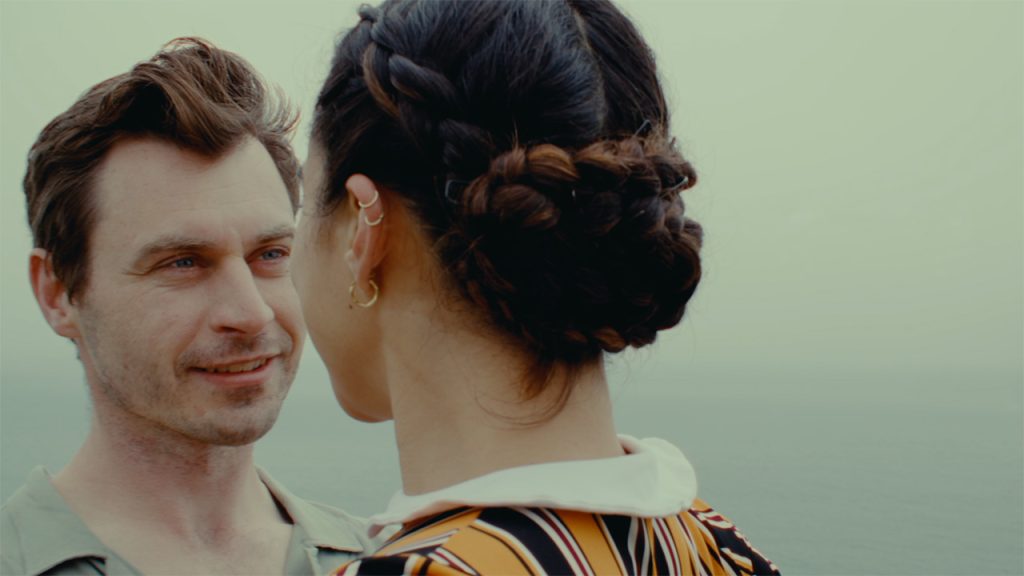 A man smiles at a person in front of him, whose back is to the camera and their hair intricately braided into a bun. The ocean fades into the gray sky in the distance.
