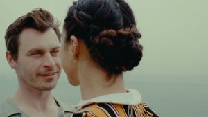 A man smiles at a person in front of him, whose back is to the camera and their hair intricately braided into a bun. The ocean fades into the gray sky in the distance.