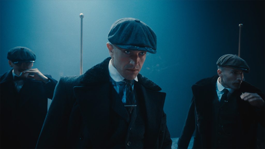 In dim, blue light, a man in a 1910s style hat and suit looks into the distance past the low brim of his hat. His face is intense and angry. Two men stand behind him, looking in the same direction.