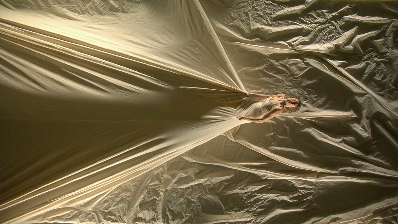 Tall bird's eye view of a person wearing a beige colored dress that extends all the way to the edges of the frame on all sides of the image. They lean back and hold the dress, pulling it taut.