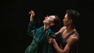 Against a black backdrop, a woman wearing a dark teal shirt leans backwards, one hand raised. Behind her, a man grips her arm while placing his other hand by her face.